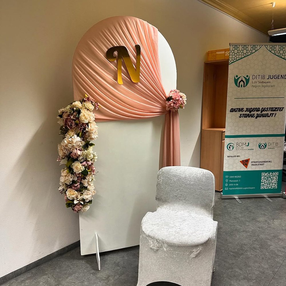 Fotowand mit rosa Stoff, Blumengestecken und einem Monogramm „N“, ergänzt durch einen weißen Stuhl.